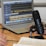 A laptop displaying audio editing software is on a wooden table. Nearby is a large microphone mounted on a stand, and the hands of two people who appear to be talking or recording. An open book is also visible, suggesting note-taking or script reading.