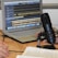 A laptop displaying audio editing software is on a wooden table. Nearby is a large microphone mounted on a stand, and the hands of two people who appear to be talking or recording. An open book is also visible, suggesting note-taking or script reading.