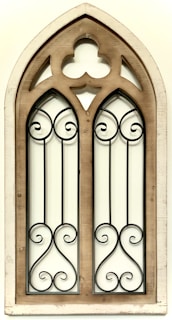 A wooden-framed gothic architectural window with intricate wrought iron detailing. The frame is slightly weathered, adding a vintage aesthetic, and the symmetrical iron patterns create an ornate look.