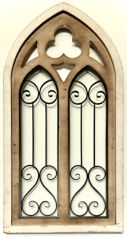 A wooden-framed gothic architectural window with intricate wrought iron detailing. The frame is slightly weathered, adding a vintage aesthetic, and the symmetrical iron patterns create an ornate look.