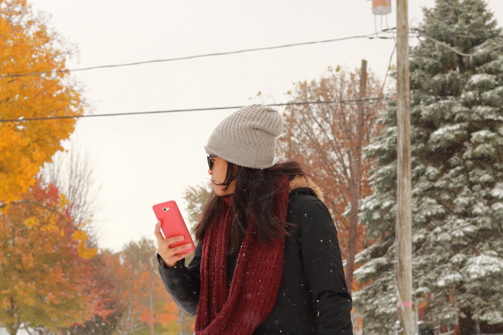 woman in black jacket and white knit cap holding red smartphone