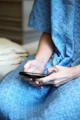 A person wearing a blue hospital gown is seated and using a smartphone. The gown has a checkered pattern with small designs. The focus is on the hands holding the mobile device.