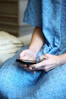A person wearing a blue hospital gown is seated and using a smartphone. The gown has a checkered pattern with small designs. The focus is on the hands holding the mobile device.