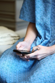 A person wearing a blue hospital gown is seated and using a smartphone. The gown has a checkered pattern with small designs. The focus is on the hands holding the mobile device.