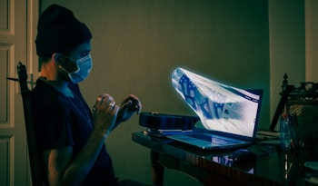 A person wearing a mask and beanie sits at a desk, interacting with a laptop. The laptop screen appears distorted with a surreal, three-dimensional image projected outward. The room is dimly lit, creating a mysterious atmosphere.