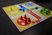 A board game setup with a colorful design, featuring four quadrants in red, yellow, green, and blue. Each quadrant has pieces matching its color, arranged in circular start areas. The board includes a pathway with directional arrows and spaces for movement. Bright plastic pieces are positioned in their respective starting zones and along the path.