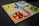 A board game setup with a colorful design, featuring four quadrants in red, yellow, green, and blue. Each quadrant has pieces matching its color, arranged in circular start areas. The board includes a pathway with directional arrows and spaces for movement. Bright plastic pieces are positioned in their respective starting zones and along the path.