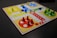 A board game setup with a colorful design, featuring four quadrants in red, yellow, green, and blue. Each quadrant has pieces matching its color, arranged in circular start areas. The board includes a pathway with directional arrows and spaces for movement. Bright plastic pieces are positioned in their respective starting zones and along the path.
