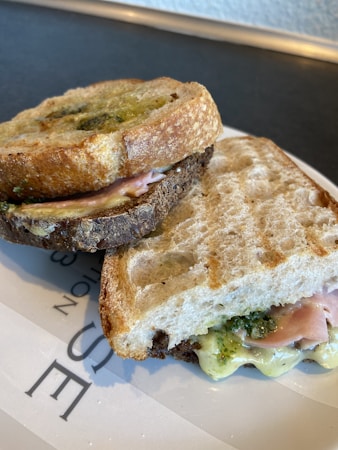 A sandwich featuring crispy, toasted bread with a golden-brown crust. Inside, slices of ham, melted cheese, and a spread of green pesto are visible. The sandwich is cut into halves and placed on a white plate with some text partially visible.