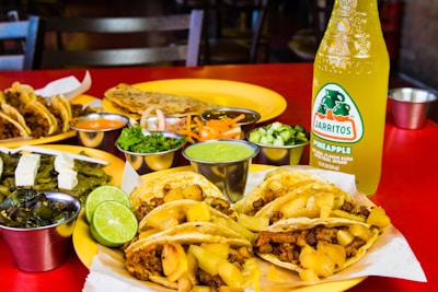 A vibrant assortment of Mexican dishes is arranged on a red table. There are tacos filled with meat and pineapple chunks, accompanied by various sides including lime wedges, grilled vegetables, and small containers of sauces and chopped herbs. In the background, additional tacos, a quesadilla, and a bottle of pineapple-flavored Jarritos soda are visible.
