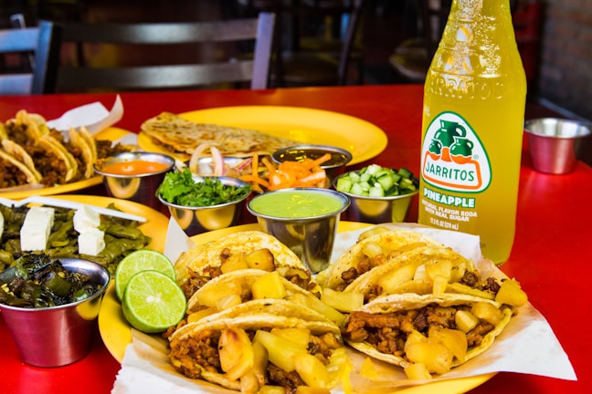 A vibrant assortment of Mexican dishes is arranged on a red table. There are tacos filled with meat and pineapple chunks, accompanied by various sides including lime wedges, grilled vegetables, and small containers of sauces and chopped herbs. In the background, additional tacos, a quesadilla, and a bottle of pineapple-flavored Jarritos soda are visible.