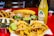 A vibrant assortment of Mexican dishes is arranged on a red table. There are tacos filled with meat and pineapple chunks, accompanied by various sides including lime wedges, grilled vegetables, and small containers of sauces and chopped herbs. In the background, additional tacos, a quesadilla, and a bottle of pineapple-flavored Jarritos soda are visible.