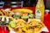 A vibrant assortment of Mexican dishes is arranged on a red table. There are tacos filled with meat and pineapple chunks, accompanied by various sides including lime wedges, grilled vegetables, and small containers of sauces and chopped herbs. In the background, additional tacos, a quesadilla, and a bottle of pineapple-flavored Jarritos soda are visible.