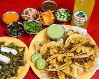 A colorful spread of Mexican food featuring several tacos filled with seasoned meat and pineapple on a yellow plate. Accompanying the tacos are lime wedges and a green sauce. Surrounding the main dish are bowls containing various garnishes such as chopped cilantro, cucumbers, sliced onions, and several sauces of different colors. A plate with sliced cactus and cheese is also visible. In the background, there is a bottle of pineapple-flavored soda.