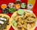 A colorful spread of Mexican food featuring several tacos filled with seasoned meat and pineapple on a yellow plate. Accompanying the tacos are lime wedges and a green sauce. Surrounding the main dish are bowls containing various garnishes such as chopped cilantro, cucumbers, sliced onions, and several sauces of different colors. A plate with sliced cactus and cheese is also visible. In the background, there is a bottle of pineapple-flavored soda.