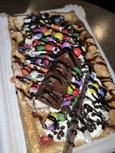 A rectangular waffle topped with whipped cream, colorful chocolate candies, chocolate syrup, and pieces of chocolate bar is displayed. The waffle is sprinkled with powdered sugar and served on a white plastic tray.