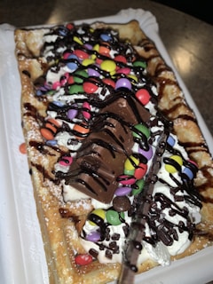 A rectangular waffle topped with whipped cream, colorful chocolate candies, chocolate syrup, and pieces of chocolate bar is displayed. The waffle is sprinkled with powdered sugar and served on a white plastic tray.