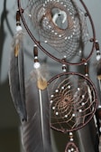 A close-up view of a dreamcatcher with intricate webbing details. The dreamcatcher is adorned with brown beads, white stones, and several large feathers which hang down elegantly. The webbing is finely crafted, featuring a circular pattern, and the feathers have a mix of gray and brown tones.