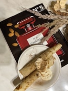 A red Chanel No. 5 perfume bottle is displayed alongside almonds and decorative dried plants on a marble surface. In the foreground, there is a plate with two rolled crepes topped with cream and sprinkled with cinnamon.