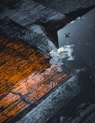 Wet pavement features a contrasting orange paint mark and a reflective puddle capturing the silhouette of an airplane flying above, along with clouds.