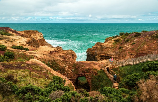 A scenic coastal view with turquoise ocean waves crashing against rugged cliffs. A wooden walkway winds through green shrubbery, providing a path for people exploring the area.