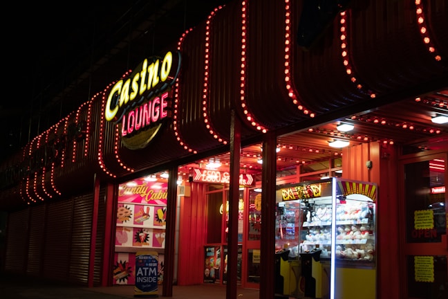 A brightly lit casino lounge entrance adorned with neon lights. The exterior features colorful signage including the words 'Casino' and 'Lounge' in vivid colors, with additional lights framing the building. Inside, a glimpse of a claw machine filled with plush toys and arcade games is visible. The area exudes an inviting yet bustling atmosphere typical of entertainment venues.