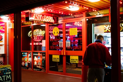 A brightly lit entrance to a casino with neon signs, including one advertising a £500 jackpot. The doors have clear signage indicating entry is restricted to those over 18. A person in a red jacket stands to the right, possibly interacting with a machine or display.