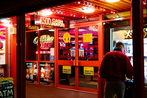 A brightly lit entrance to a casino with neon signs, including one advertising a £500 jackpot. The doors have clear signage indicating entry is restricted to those over 18. A person in a red jacket stands to the right, possibly interacting with a machine or display.