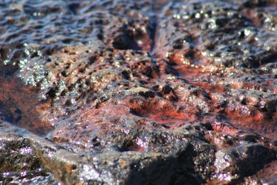 A close-up view of a rough, textured surface resembling volcanic rock or lava. The surface features shiny, reflective patches with intricate patterns and grooves, giving it a dynamic appearance.