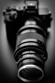 A black and white photograph of a camera with an extended lens. The image is focused on the lens with intricate details on its rings and markings, creating a sense of depth.
