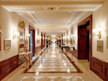 A luxurious and elegant corridor featuring a polished marble floor with an intricate pattern. Ornate gold-framed mirrors and wall sconces line the walls, which are decorated with framed artworks. The warm lighting adds to the opulent atmosphere, and decorative tables and potted plants are placed intermittently along the pathway.