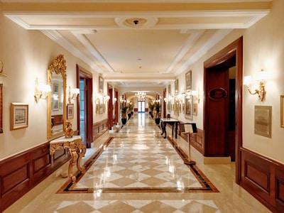 A luxurious and elegant corridor featuring a polished marble floor with an intricate pattern. Ornate gold-framed mirrors and wall sconces line the walls, which are decorated with framed artworks. The warm lighting adds to the opulent atmosphere, and decorative tables and potted plants are placed intermittently along the pathway.