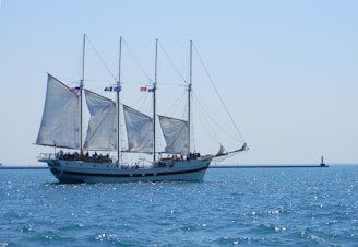 A large sailboat with three masts sailing on calm blue waters. The boat is adorned with multiple sails, and there are several people aboard the vessel, enjoying the ride. Flags are visible atop the masts, and in the background, a lighthouse stands on a breakwater.