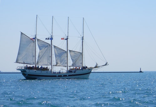 A large sailboat with three masts sailing on calm blue waters. The boat is adorned with multiple sails, and there are several people aboard the vessel, enjoying the ride. Flags are visible atop the masts, and in the background, a lighthouse stands on a breakwater.