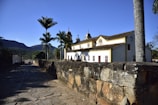 A historic colonial-style building with a white facade and yellow accents stands adjacent to a cobblestone path. Tall palm trees are present in the courtyard area beside the building, and distant hills and clear skies add to the scenic setting.