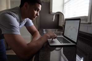 A person is focused on a laptop displaying stock market charts while standing at a kitchen counter. The room is well-lit with natural light streaming in through the window blinds.