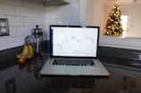A laptop displaying a stock chart is placed on a granite kitchen countertop next to a bunch of bananas and a glass French press. In the background, there is a decorated Christmas tree with lights, visible through an opening in the wall.