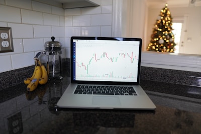 A laptop displaying a stock chart is placed on a granite kitchen countertop next to a bunch of bananas and a glass French press. In the background, there is a decorated Christmas tree with lights, visible through an opening in the wall.