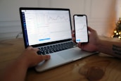 A person is sitting at a wooden table using a laptop and a smartphone. Both devices display stock charts and market data, suggesting activities related to financial analysis or trading. The room is softly lit, possibly with a Christmas tree in the background.