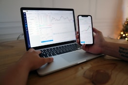 A person is sitting at a wooden table using a laptop and a smartphone. Both devices display stock charts and market data, suggesting activities related to financial analysis or trading. The room is softly lit, possibly with a Christmas tree in the background.