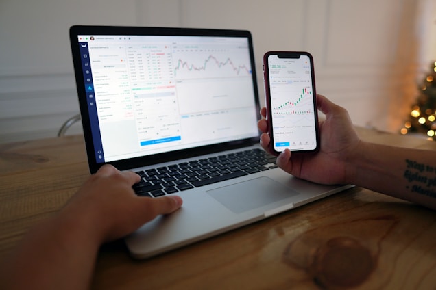 A person is sitting at a wooden table using a laptop and a smartphone. Both devices display stock charts and market data, suggesting activities related to financial analysis or trading. The room is softly lit, possibly with a Christmas tree in the background.
