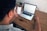 A person is sitting at a wooden table, looking at a smartphone and a laptop. Both screens display stock market charts and data. The person is holding the phone in one hand while the other hand rests on the laptop keyboard.