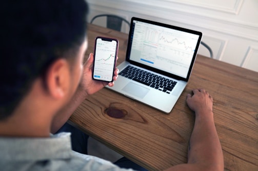 A person is sitting at a wooden table, looking at a smartphone and a laptop. Both screens display stock market charts and data. The person is holding the phone in one hand while the other hand rests on the laptop keyboard.