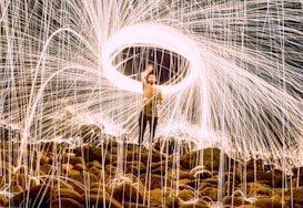 A person stands on a rocky surface, surrounded by a stunning display of long-exposure light effects creating a whirl of fiery sparks. The circular pattern of light appears like a halo above the person, with bright streaks extending in all directions, creating a dynamic and energetic visual.