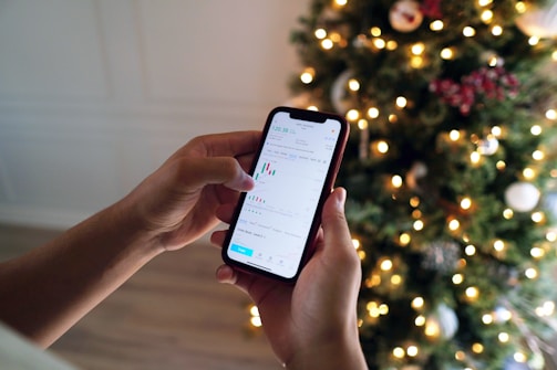 A person holding a smartphone displaying a financial app with charts and data, while standing near a decorated Christmas tree with glowing lights.