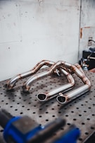Several polished metal exhaust pipes arranged on a perforated metal table, with a plain industrial wall in the background. The pipes exhibit a clean and shiny finish with weld marks visible at the joints.