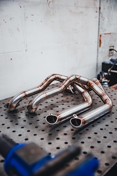 Several polished metal exhaust pipes arranged on a perforated metal table, with a plain industrial wall in the background. The pipes exhibit a clean and shiny finish with weld marks visible at the joints.