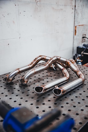 Several polished metal exhaust pipes arranged on a perforated metal table, with a plain industrial wall in the background. The pipes exhibit a clean and shiny finish with weld marks visible at the joints.