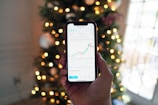 A hand holding a smartphone displaying a stock market chart with candlesticks and line graphs. In the background, there are blurred Christmas tree lights creating a festive ambiance.