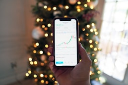 A hand holding a smartphone displaying a stock market chart with candlesticks and line graphs. In the background, there are blurred Christmas tree lights creating a festive ambiance.